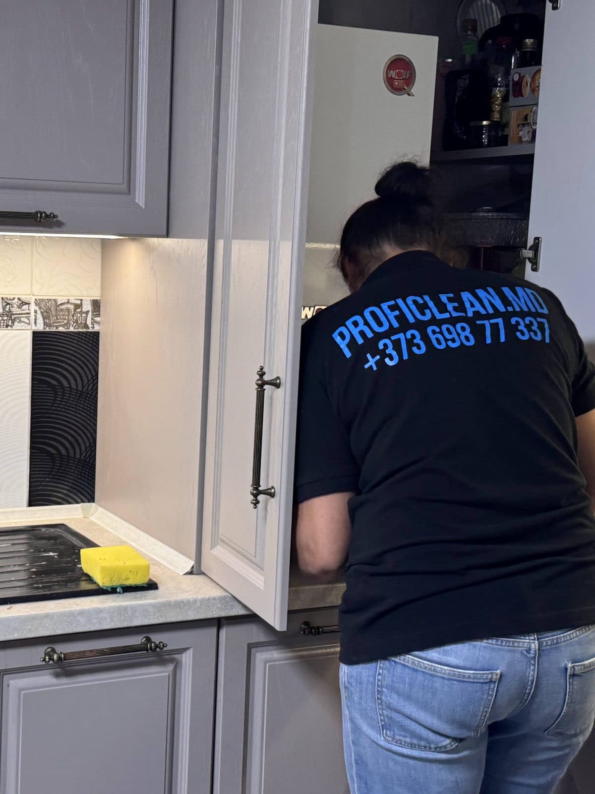 Colegă ProfiClean, în tricoul firmei cu numărul +373 698 77 337, șterge dulapurile de bucătărie pe interior